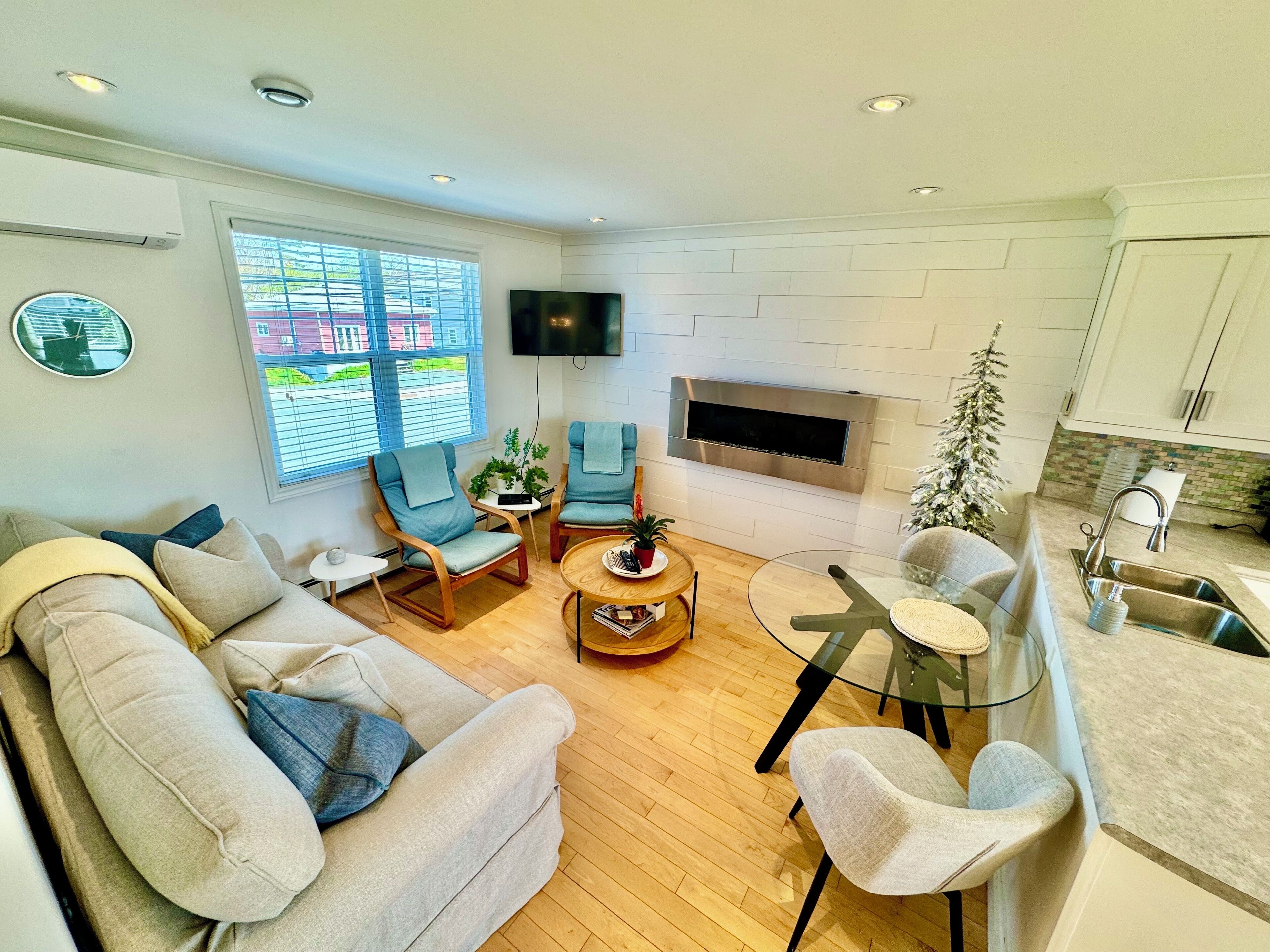 A modern living room with a sofa, two armchairs, a coffee table, and a TV. There is a fireplace and a kitchen area with a sink and countertop.