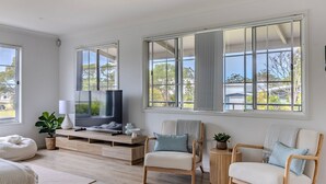 A modern living room with a large TV, wooden furniture, and large windows with views of the neighborhood. - The Sussex Beach Bach (Sussex Inlet)