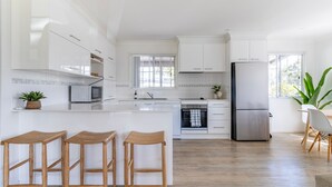 A modern kitchen with white cabinetry, a central island with stools, a microwave, oven, and refrigerator. - The Sussex Beach Bach (Sussex Inlet)