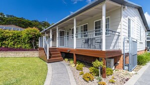 A single-story house with a covered porch, a small front yard with a variety of plants, and a solar panel array in the background. - The Sussex Beach Bach (Sussex Inlet)