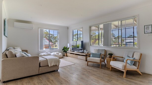 A spacious living room with a sofa, chairs, a TV, and large windows offering views of the outside. - The Sussex Beach Bach (Sussex Inlet)
