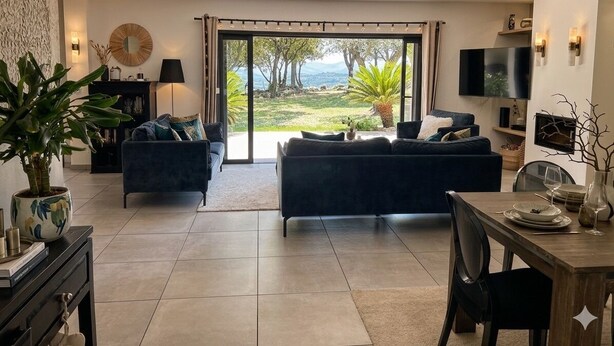 Una sala de estar moderna con un sofá, una mesa de comedor y un televisor. Hay un ventanal con vistas a zonas verdes y una montaña.