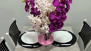 A table set with white plates, black placemats, and black cutlery. A vase with purple and white flowers is placed in the center.