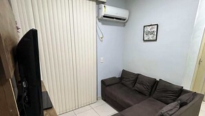 A living room with a sectional sofa, a TV on a wooden stand, a white air conditioner, and a framed picture on the wall.