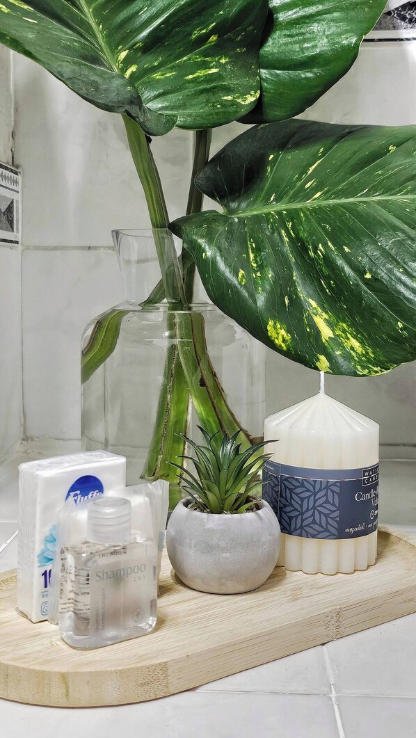 A bathroom vanity with a large green plant, a glass vase, a small potted plant, and toiletries including a shampoo bottle and candles.