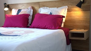 Ein Bett mit weißer Bettwäsche und zwei roten Kissen, ein hölzernes Kopfteil, ein Nachttisch mit Lampe sowie eine Wandleuchte.