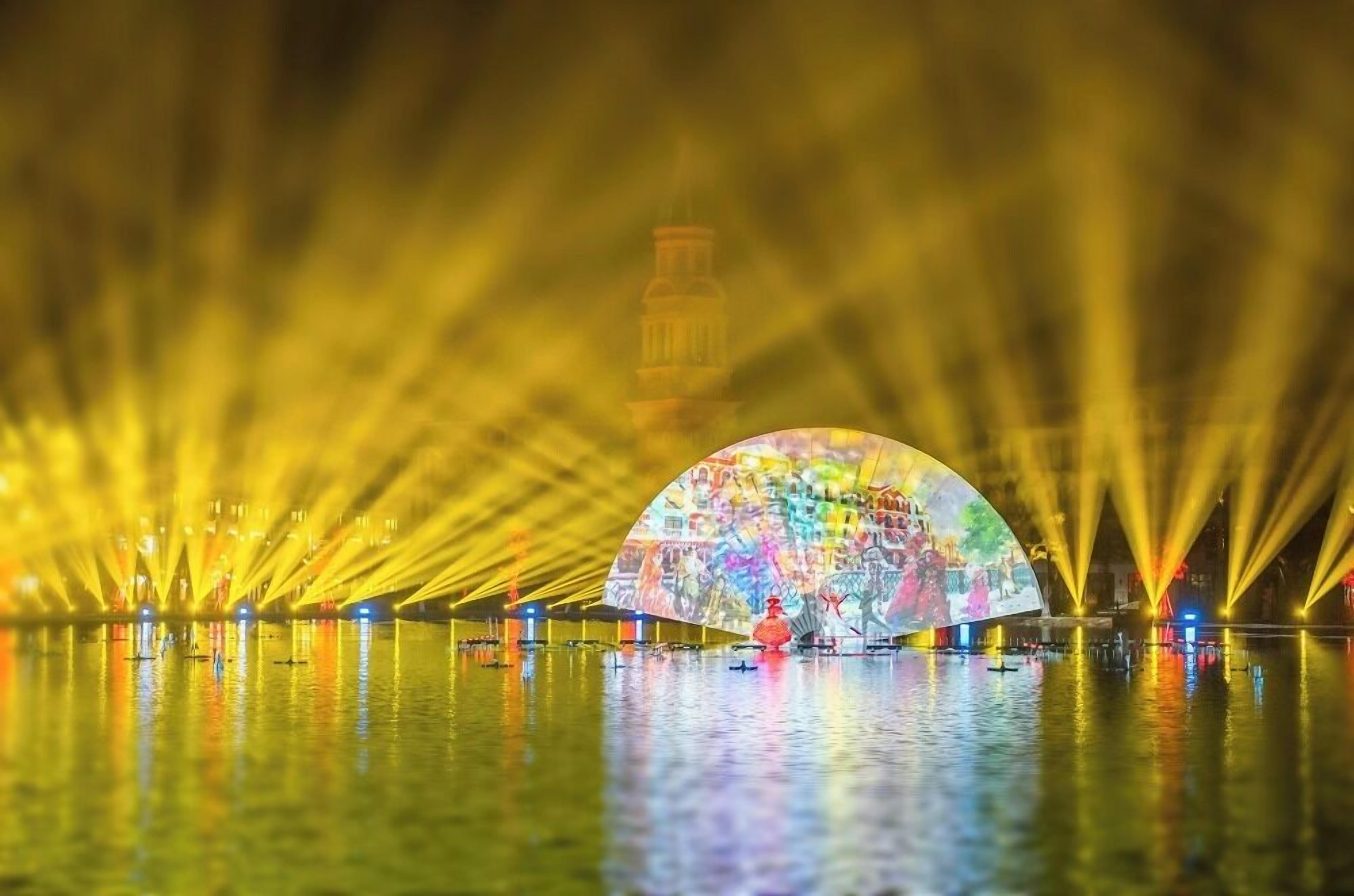 Una gran estructura en forma de cúpula iluminada, con un mural colorido en su superficie, rodeada de agua y reflejos de luces.