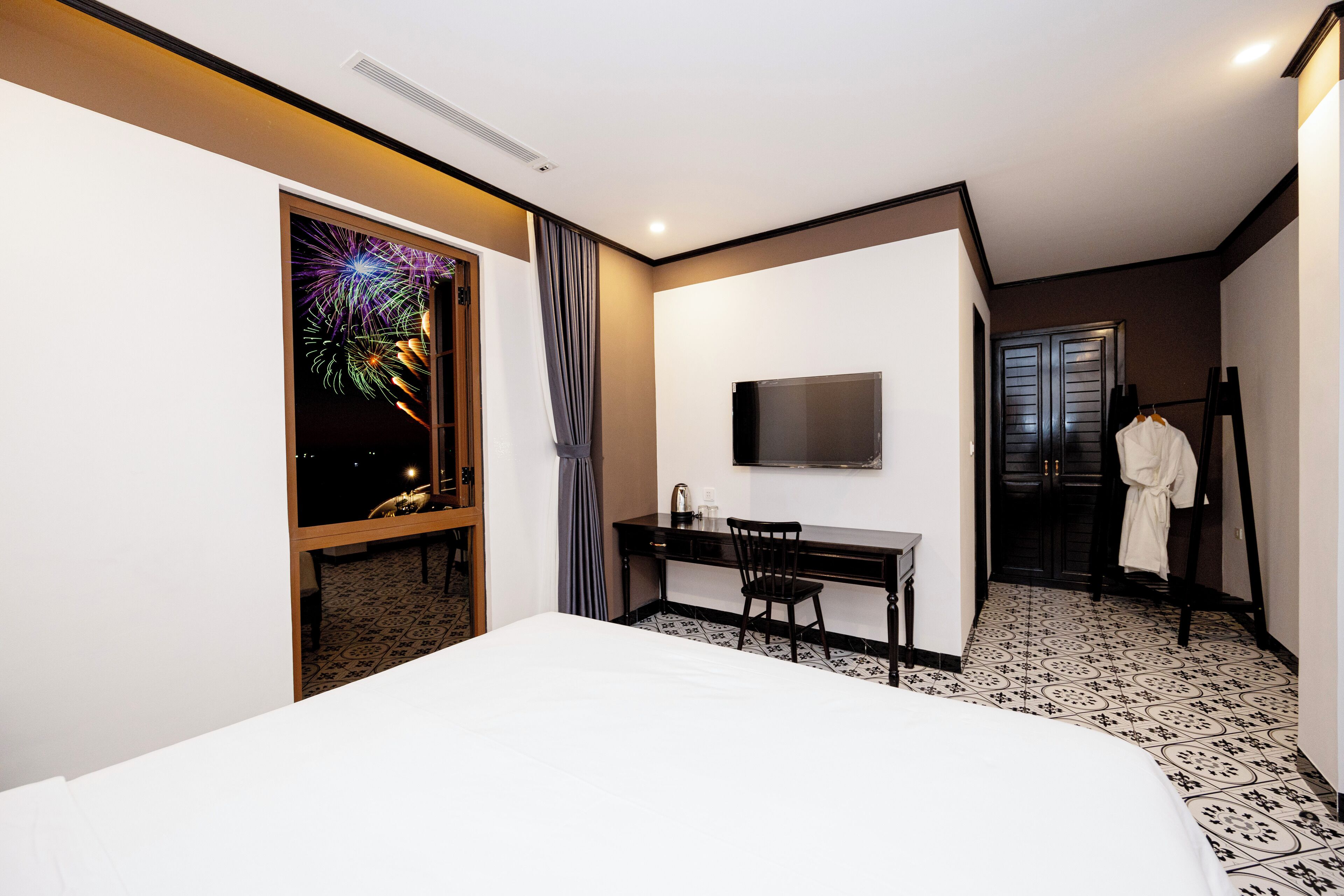 A hotel room with a bed, a desk, a chair, a TV mounted on the wall, and a robe hanging on a rack.