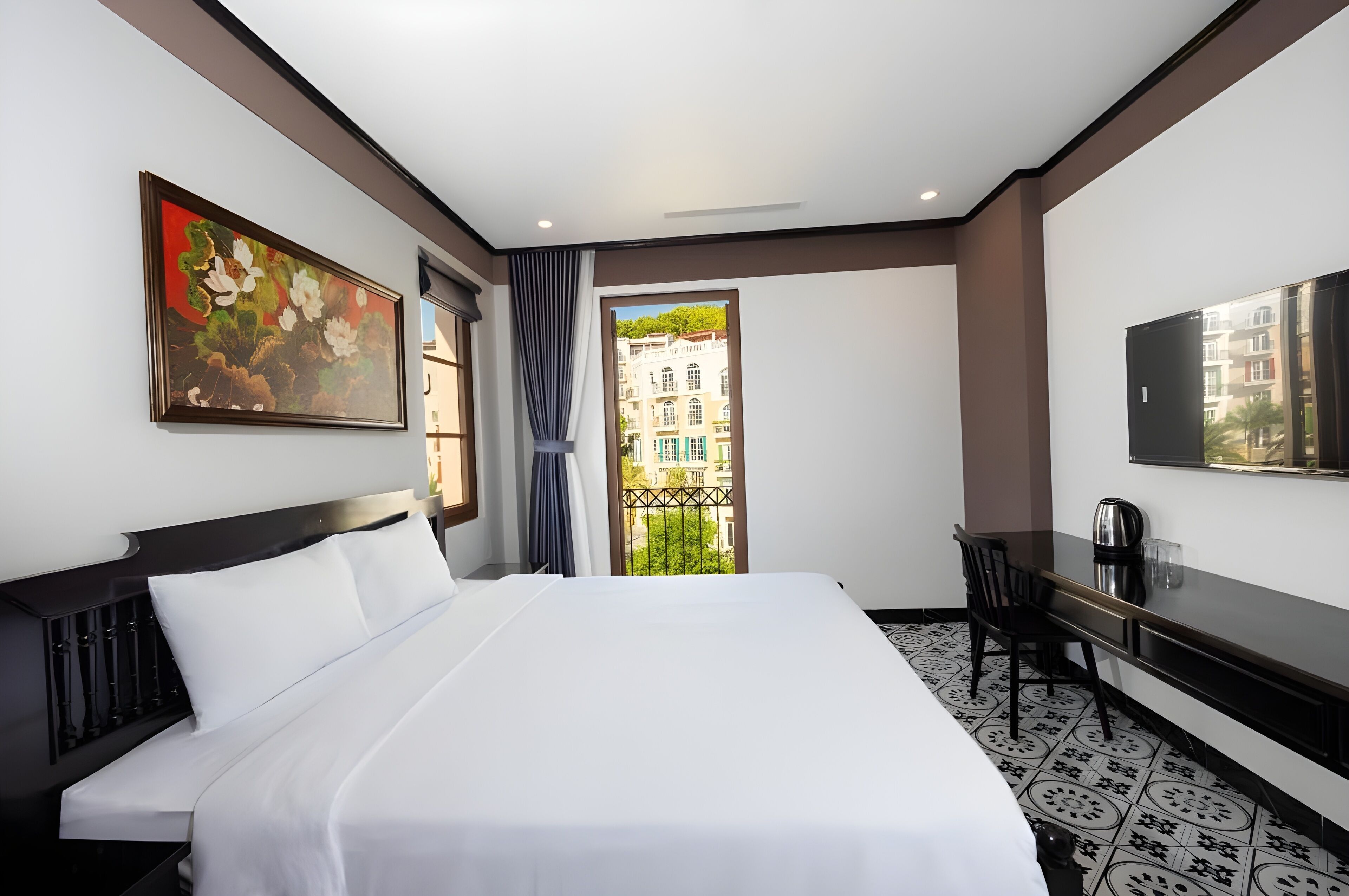 A hotel room with a large bed, a desk, a chair, a painting, and a view of buildings through a window.