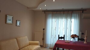 Een woonkamer met een beige bank, een eettafel met een rood tafelkleed en een raam met lichte gordijnen.