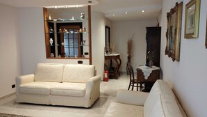 Een woonkamer met een beige bank, een kamerbreed tapijt en een glazen vitrinekast met decoratieve objecten.