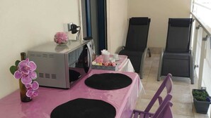 Een klein terras met een roze tafel, twee zwarte stoelen, een paarse stoel, een kleine potplant en een raam met uitzicht.