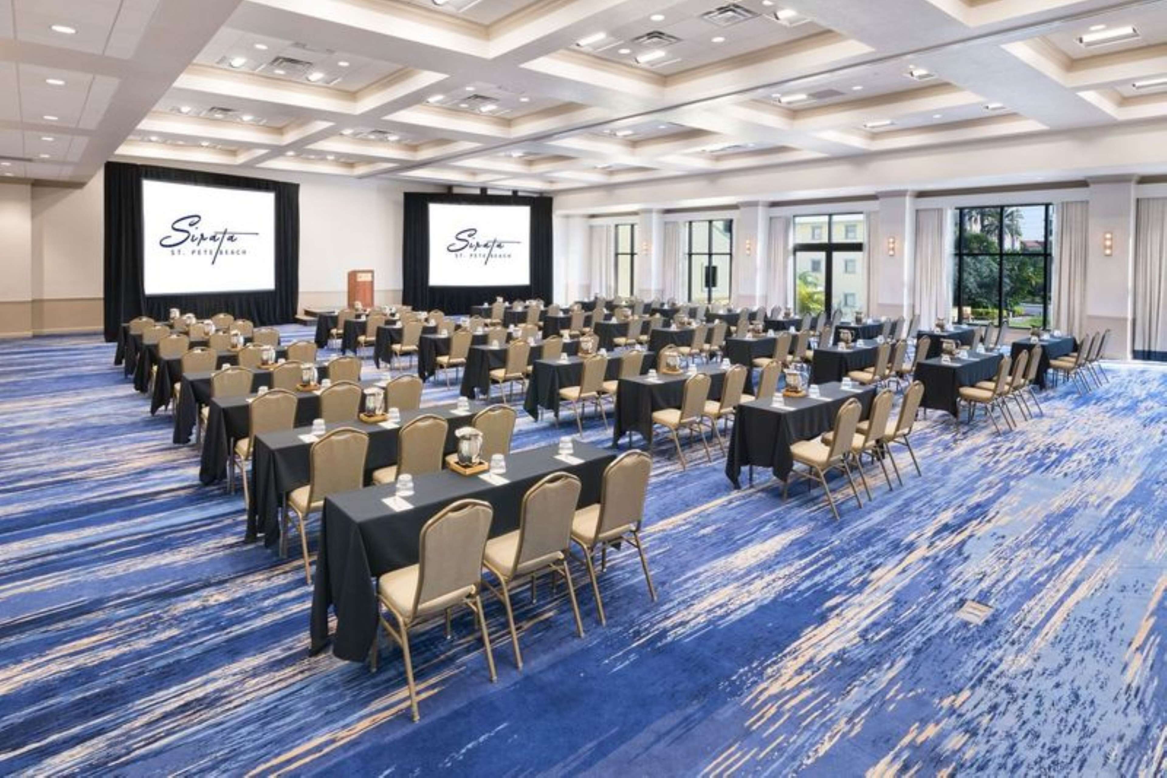 Un salón de conferencias con filas de mesas y sillas, pantallas grandes y suelo cubierto de moqueta azul.
