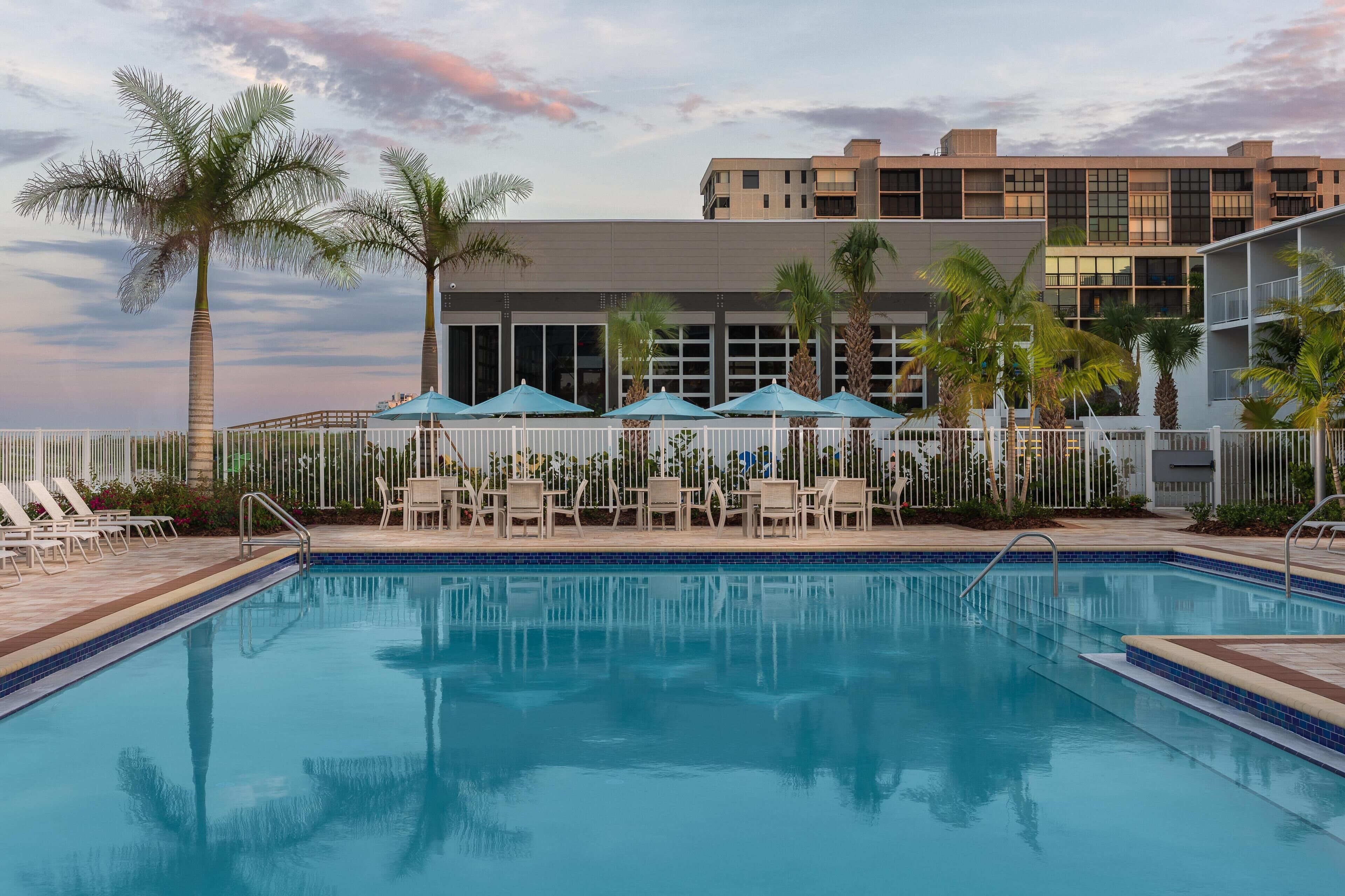 A swimming pool with lounge chairs and umbrellas, surrounded by palm trees and a building with large windows.