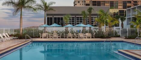 Una piscina con sillas de descanso y sombrillas, rodeada de palmeras y un edificio con amplias ventanas.