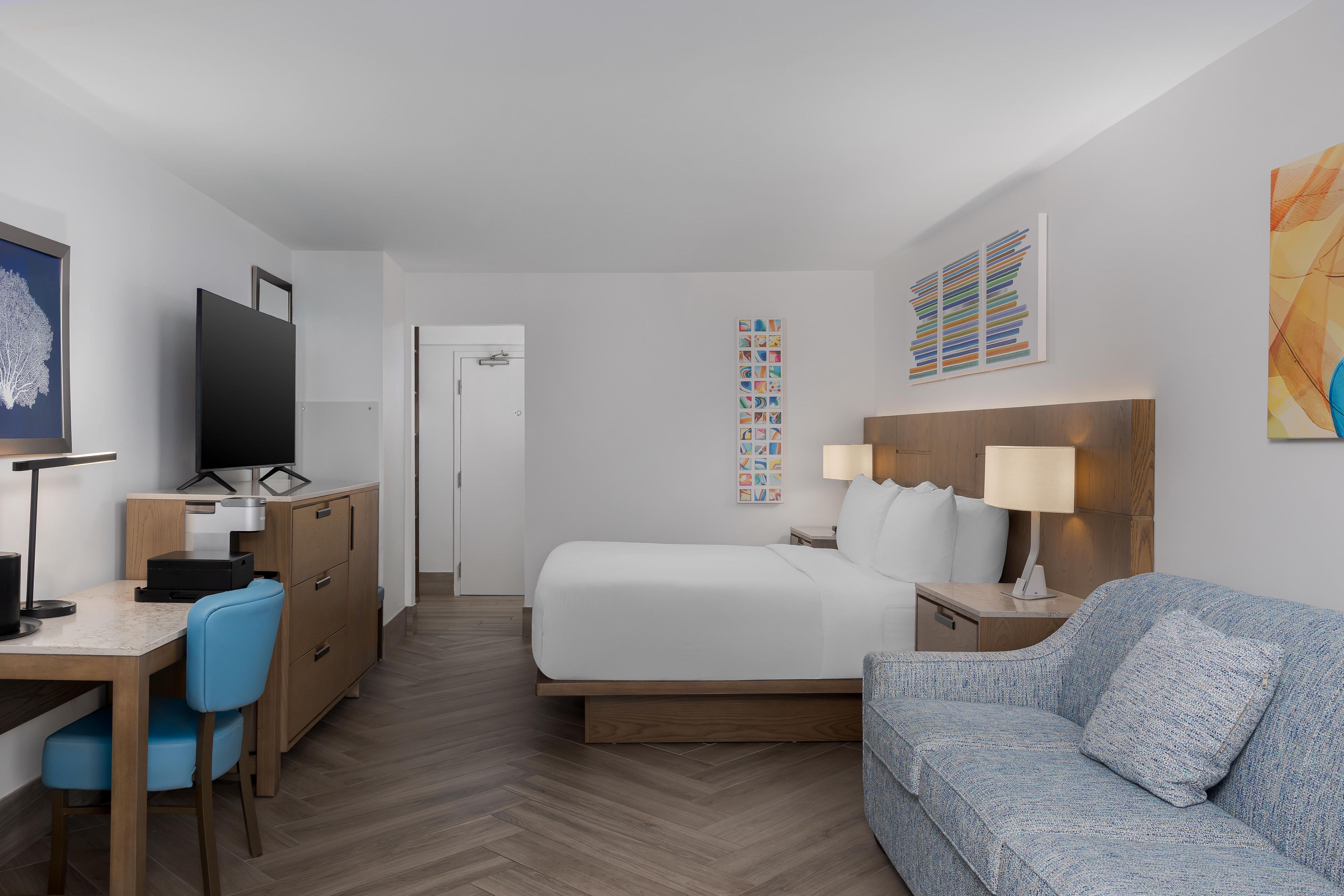 Una habitación de hotel con cama, escritorio con computadora, una silla, sofá y obras de arte en las paredes.