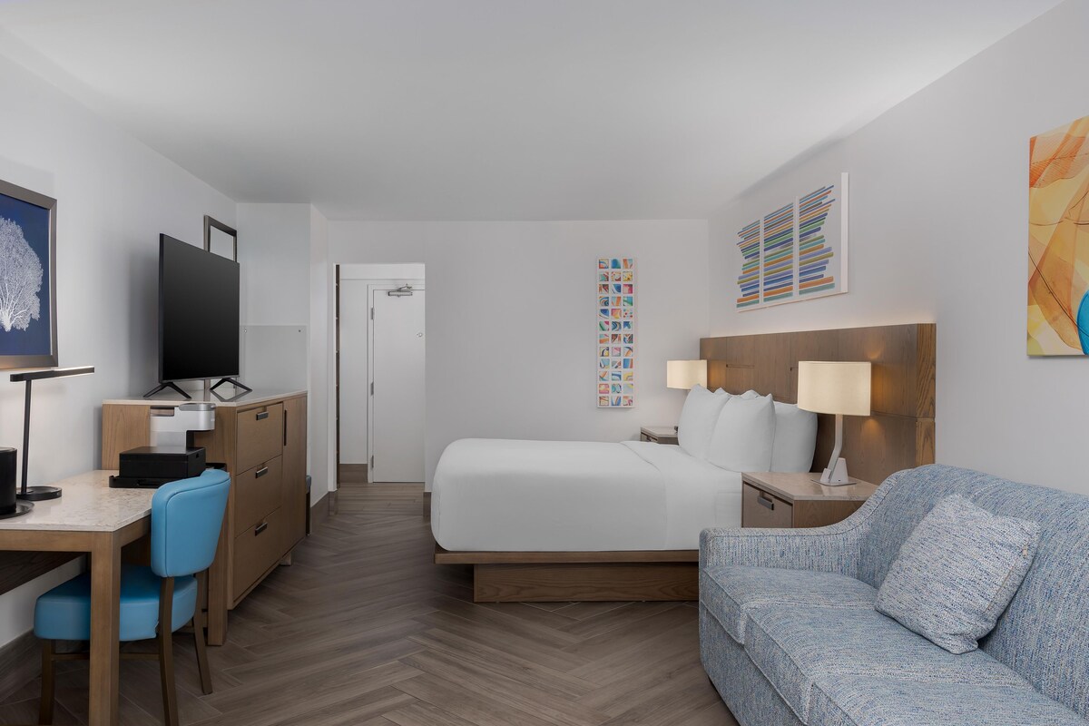 Una habitación de hotel con cama, escritorio con computadora, una silla, sofá y obras de arte en las paredes.