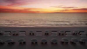 Una playa con filas de sillas de descanso y sombrillas a rayas, frente al océano al atardecer.