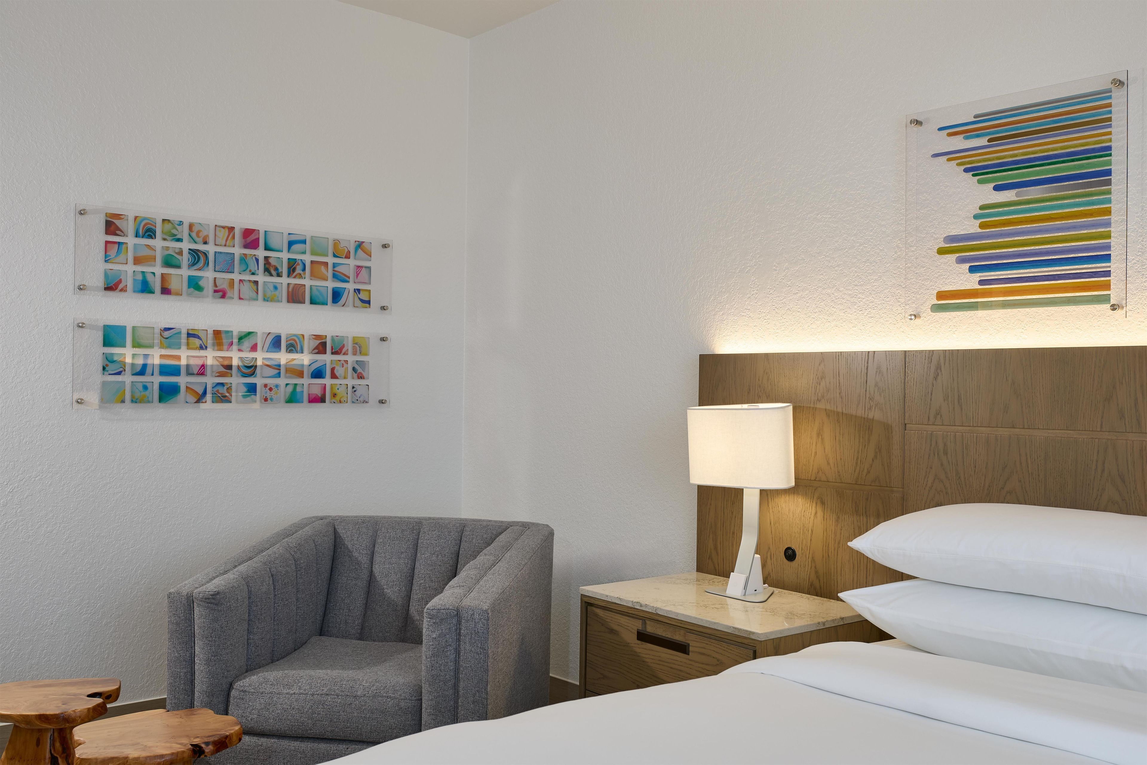 Een hotelkamer met een grijze fauteuil, een nachtkastje met een lamp, een bed met wit beddengoed, en twee kunstwerken aan de muur.