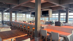 Un restaurante con zona de bar, mesas y grandes ventanales con vistas al mar.
