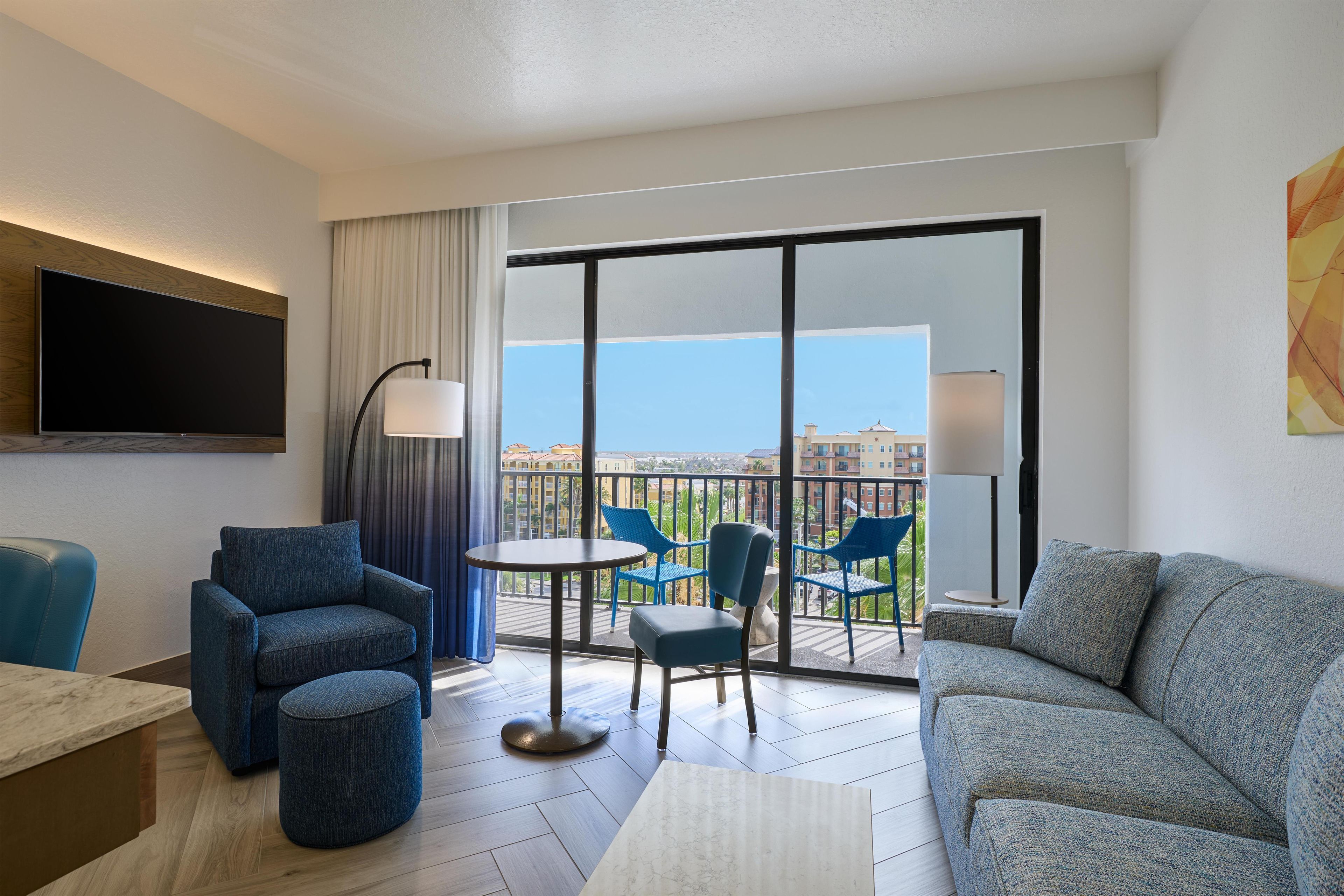 A hotel room with a balcony, a flat-screen TV, a sofa, a small table, and two chairs.
