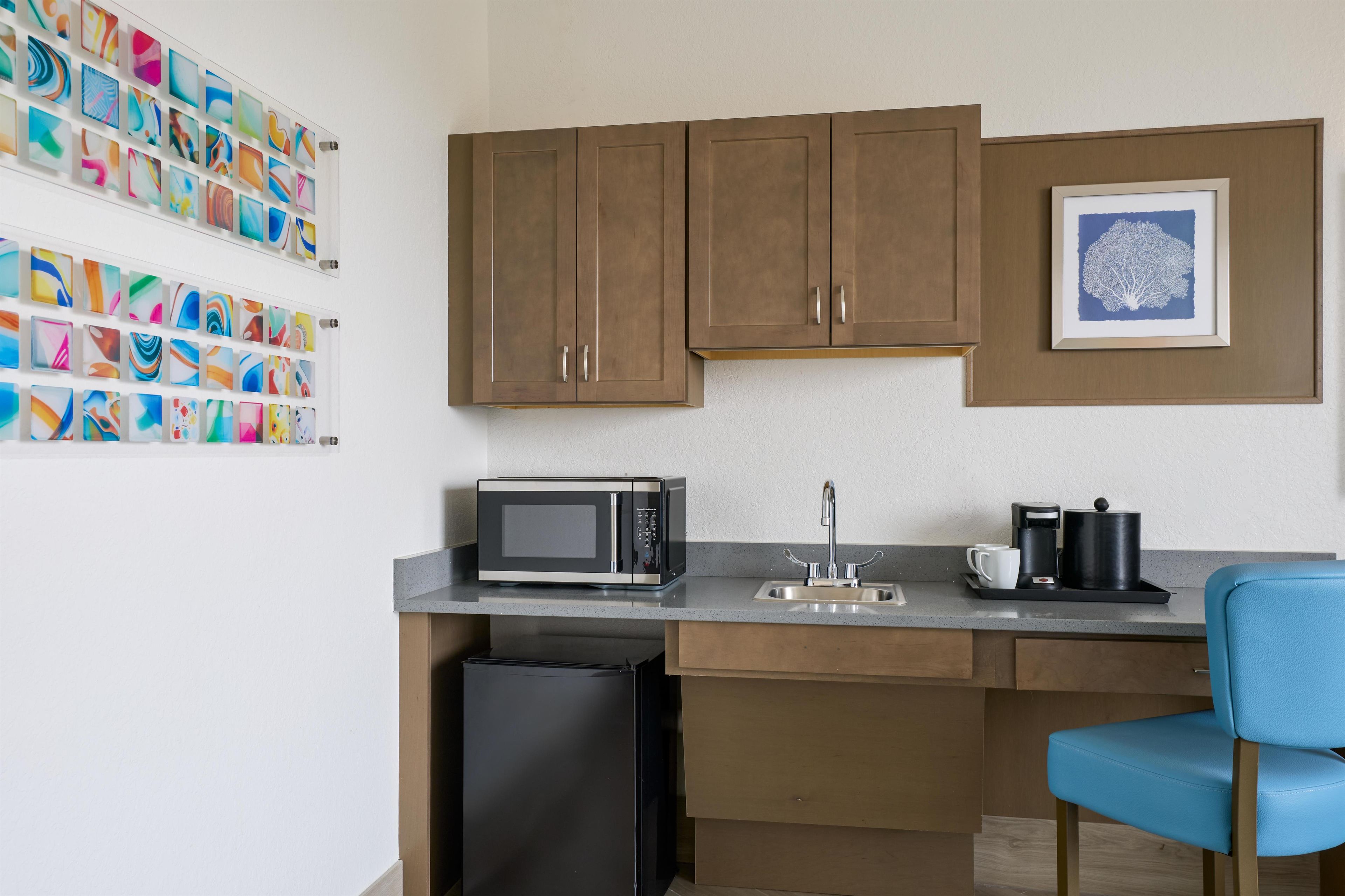 A compact kitchenette with a microwave, coffee maker, and sink. There is a trash bin and a blue chair.