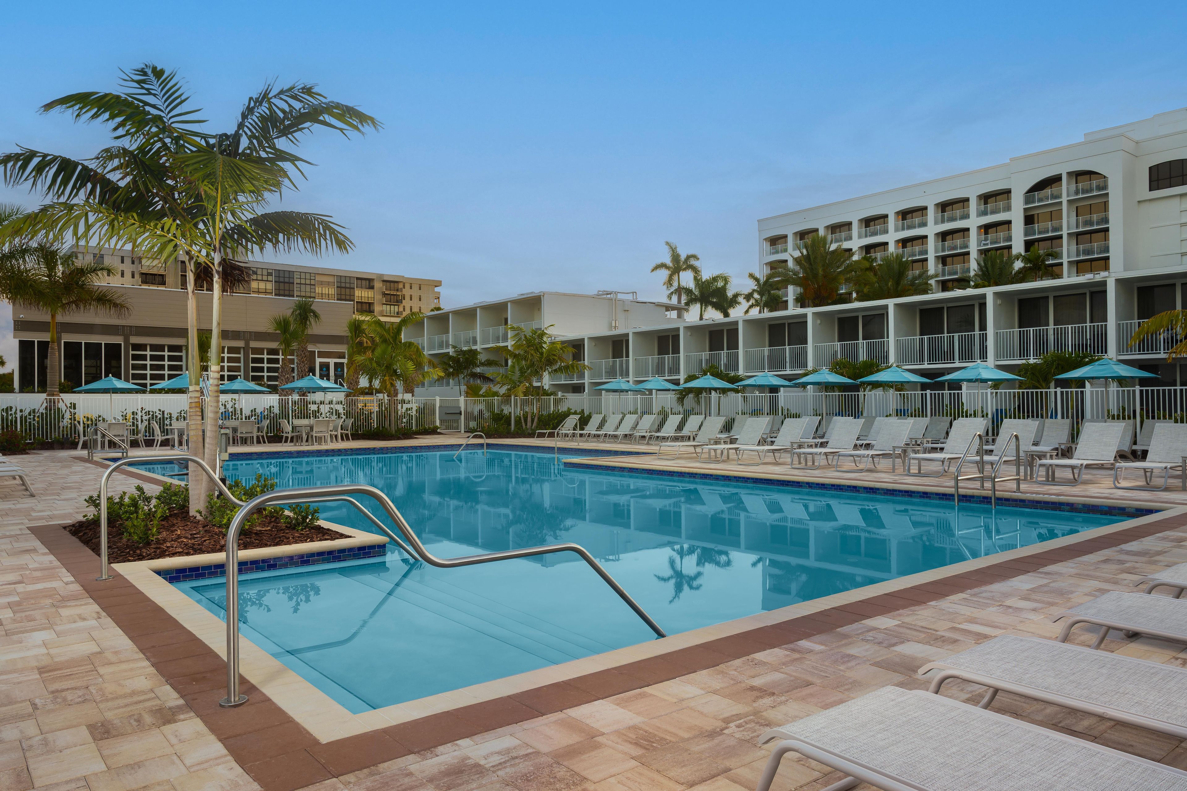 A swimming pool with lounge chairs and umbrellas, surrounded by palm trees and a hotel building.