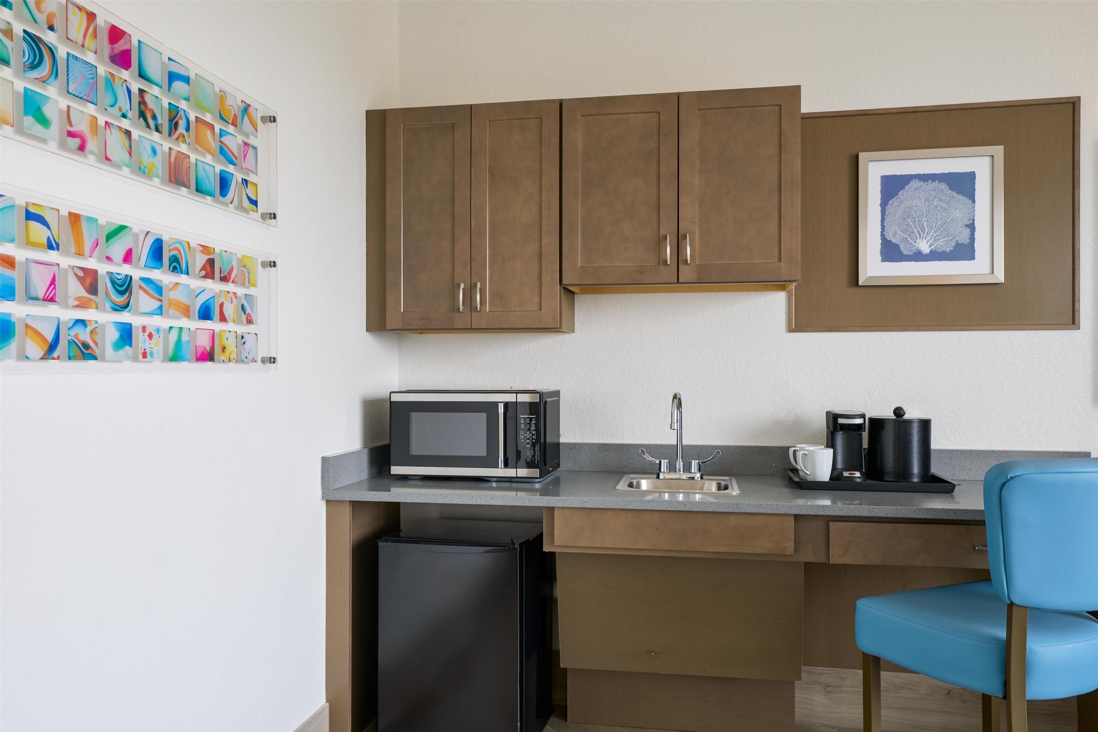 A compact kitchen with a microwave, sink, and coffee maker. There is a trash bin and a blue chair.