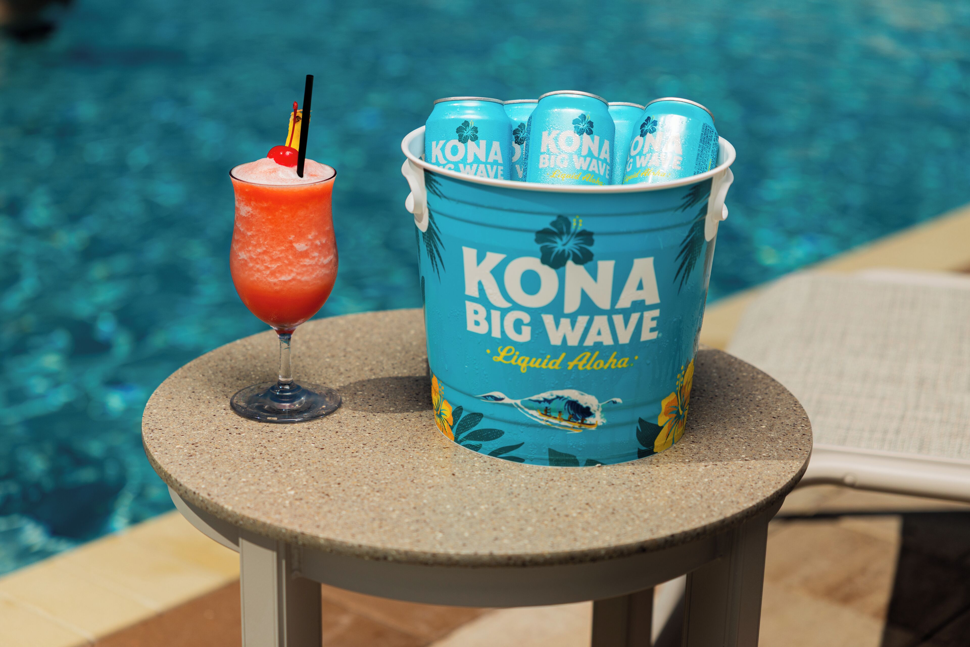 Ein Eimer KONA Big Wave Bierdosen und ein Glas rotes Getränk mit Trinkhalm auf einem Tisch am Pool.