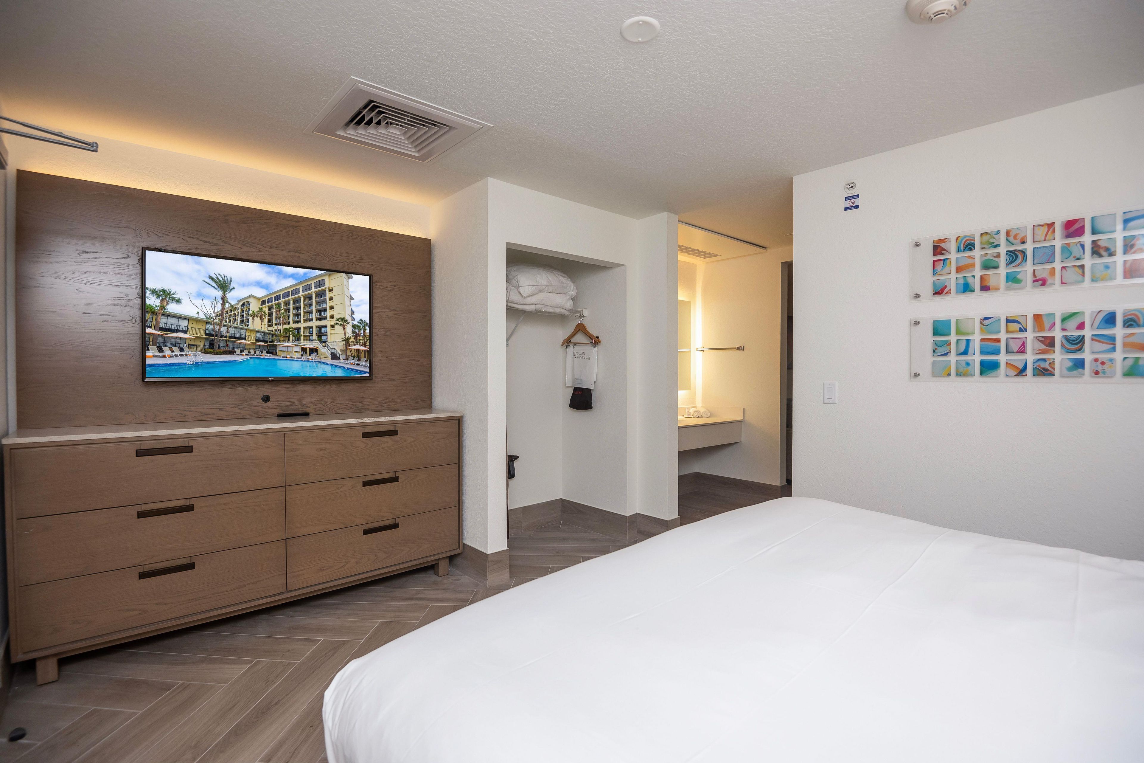 Quarto de hotel com uma cama grande, cómoda de madeira, televisão presa à parede e banheiro visível através de uma porta aberta.