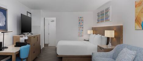A hotel room with a bed, a desk with a computer, a chair, a sofa, and artwork on the walls.