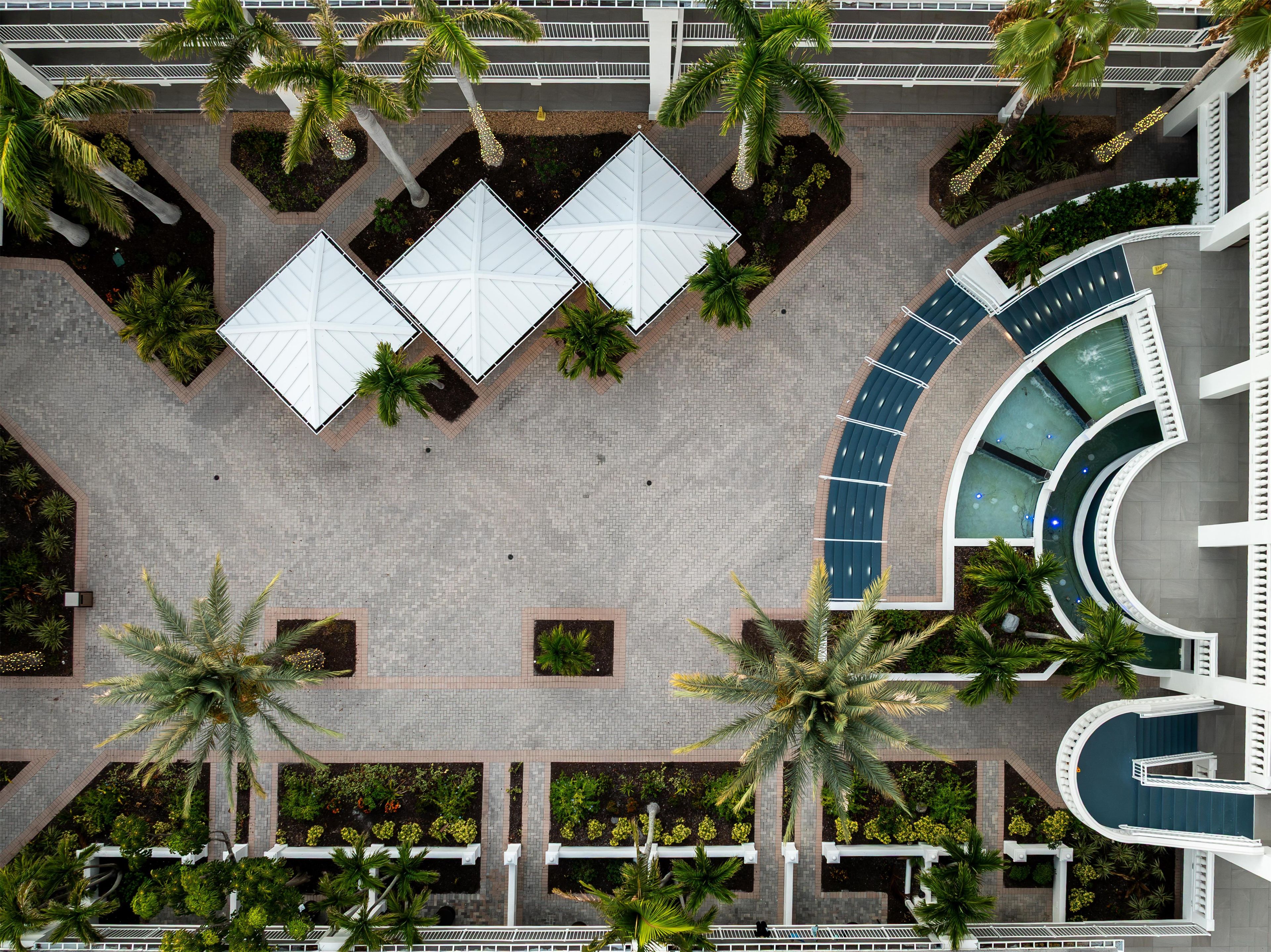 An aerial view of a hotel courtyard with palm trees, white umbrellas, and a curved pool area.