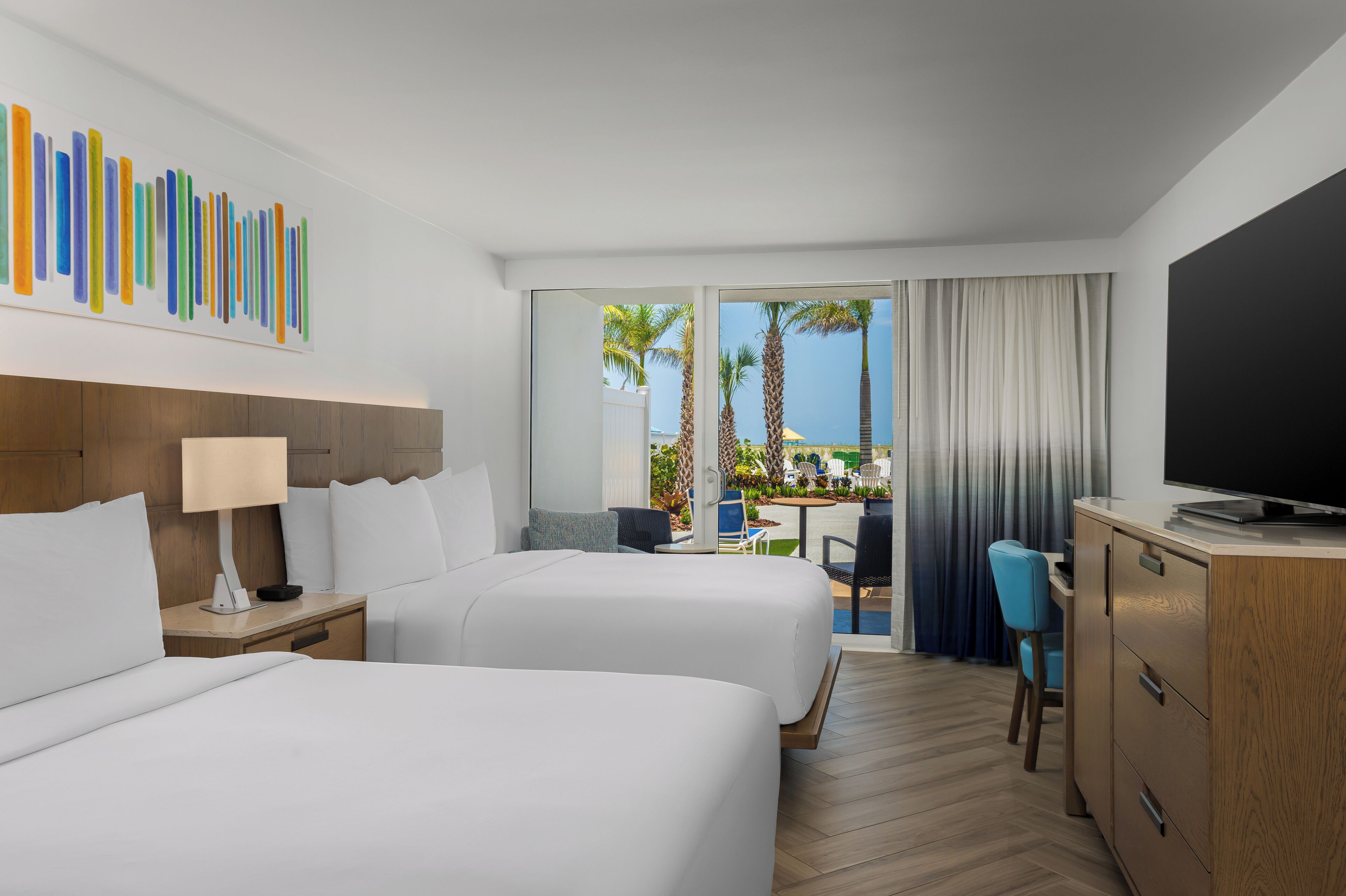 A hotel room with two beds, a desk, a TV, and a view of palm trees and a pool.