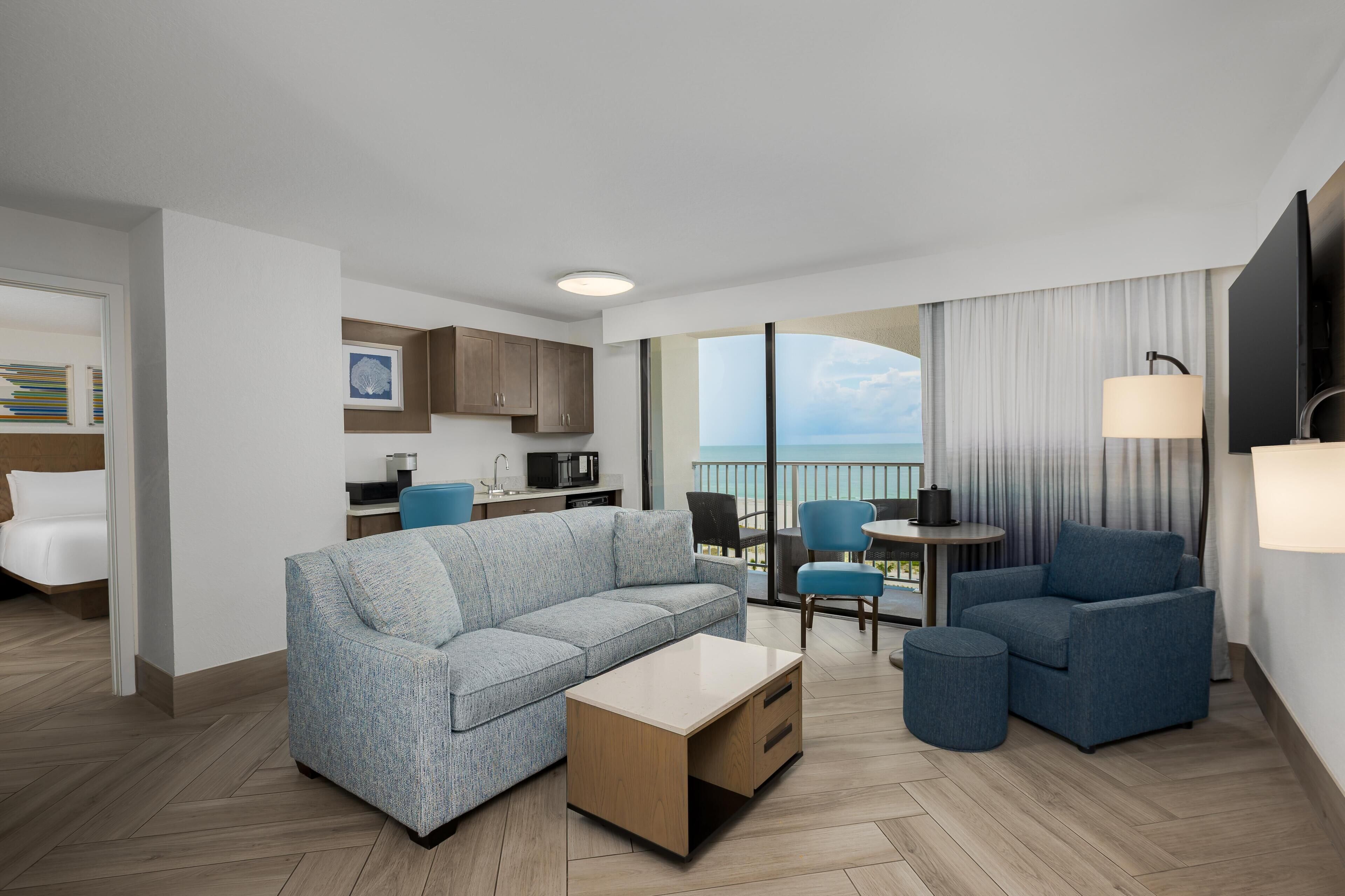 A modern hotel room with a sofa, a coffee table, a dining table, and a balcony with a view.