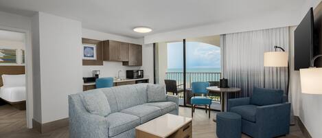 A modern hotel room with a sofa, a coffee table, a dining table, and a balcony with a view.