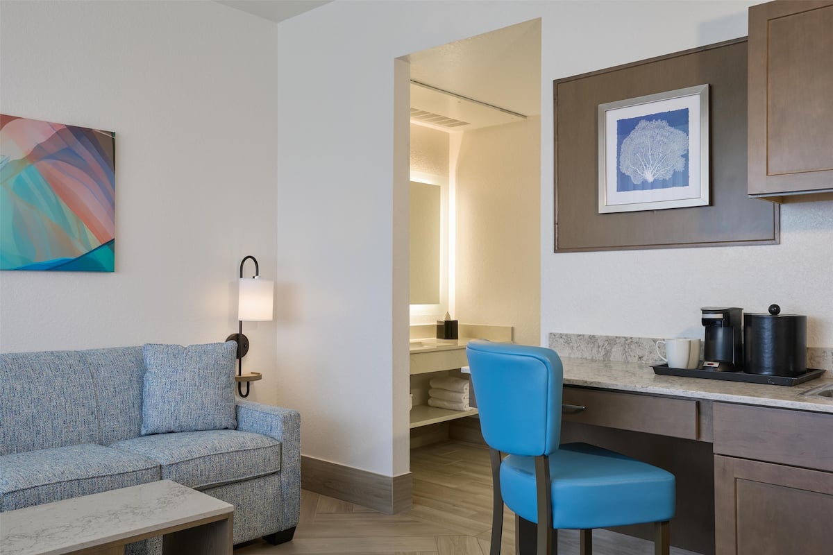 Una habitación de hotel moderna con sofá, una silla azul, una mesita, una zona de cocina con cafetera y un cuadro en la pared.