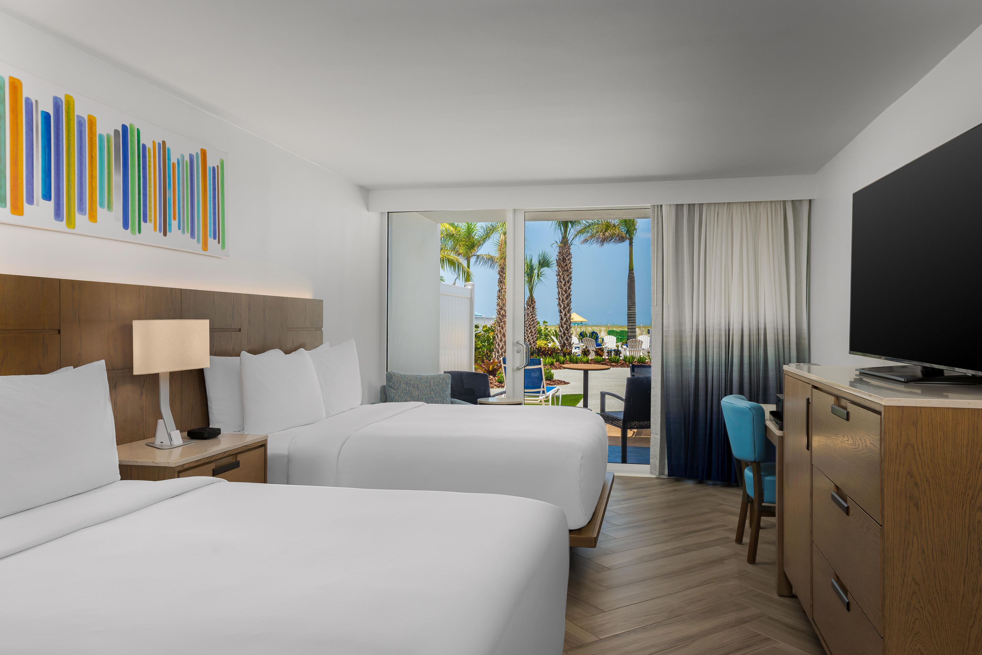 A hotel room with two beds, a desk, a television, and a view of palm trees and a pool.