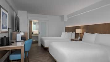 Quarto de hotel com duas camas, uma escrivaninha com cafeteira, uma cadeira e um banheiro visível por uma porta aberta.
