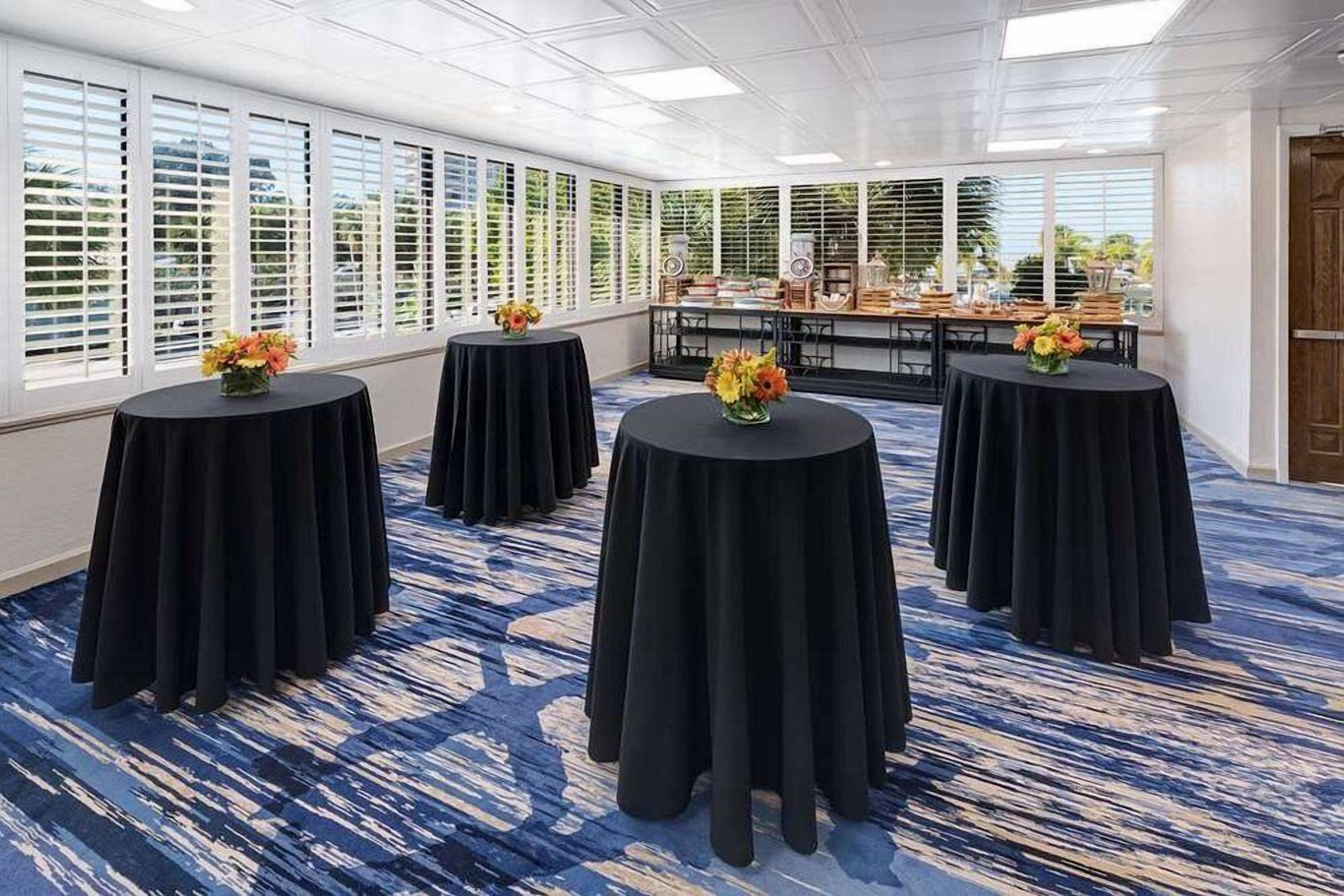 Une salle de conférence avec quatre tables rondes, chacune recouverte d’une nappe noire et agrémentée d’un vase de fleurs. La pièce est dotée de grandes fenêtres équipées de stores, d’un sol recouvert de moquette et, au fond, d’une table en bois.