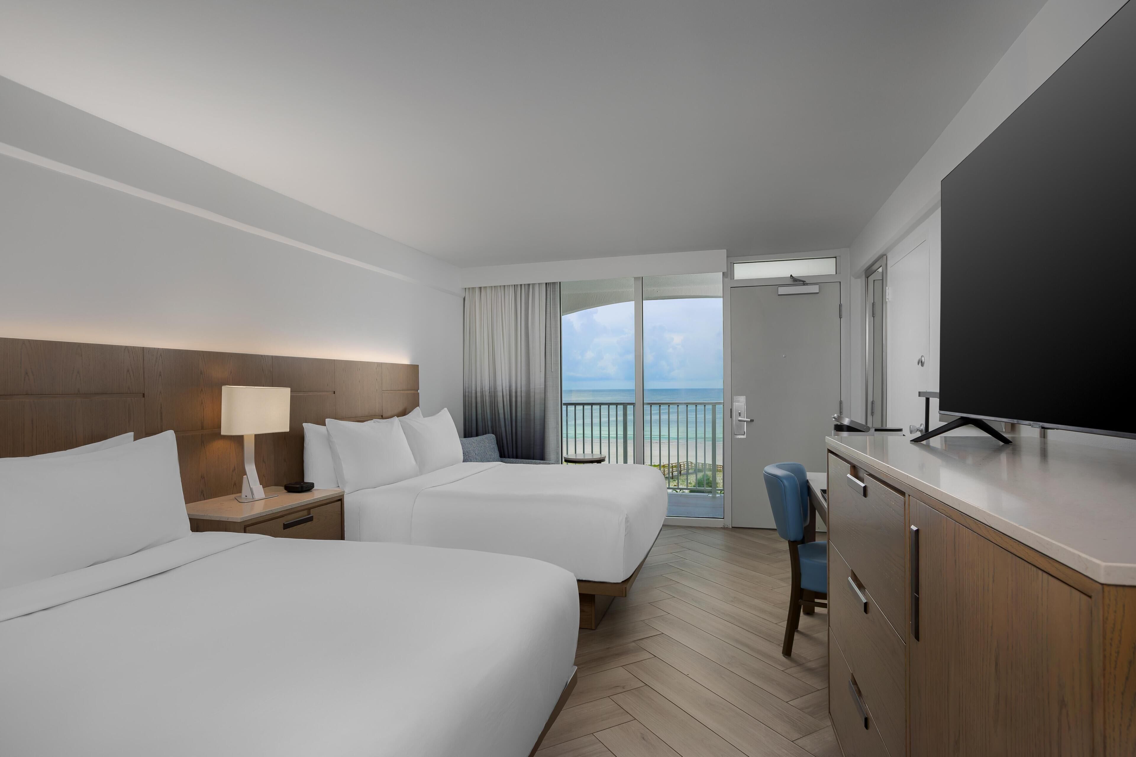 A hotel room with two beds, a TV, a desk, and a view of the ocean.