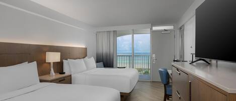 A hotel room with two beds, a TV, a desk, and a view of the ocean.