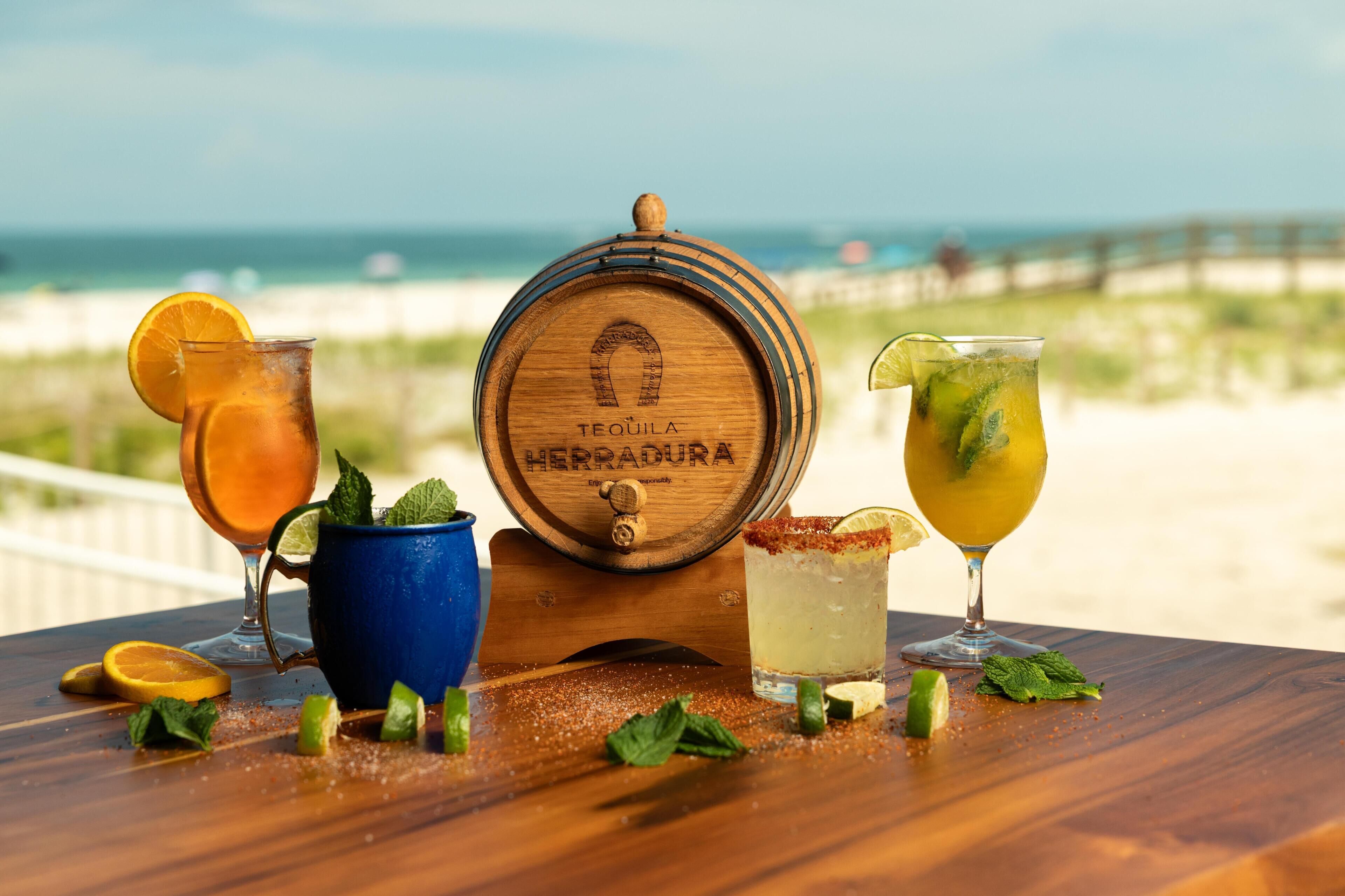 A wooden barrel with 'TEQUILA HERRADURA' engraved on it, surrounded by three drinks with garnishes, on a wooden table with a beach view in the background.