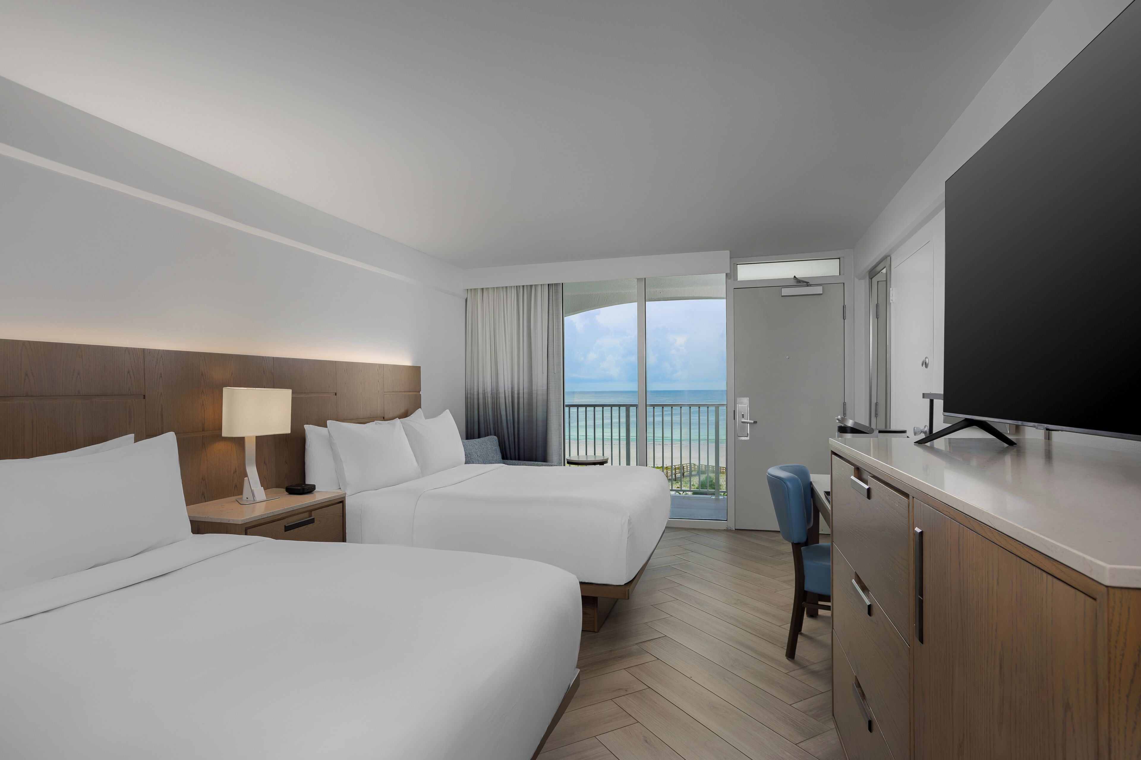 A hotel room with two beds, a TV, a desk, and a view of the ocean.