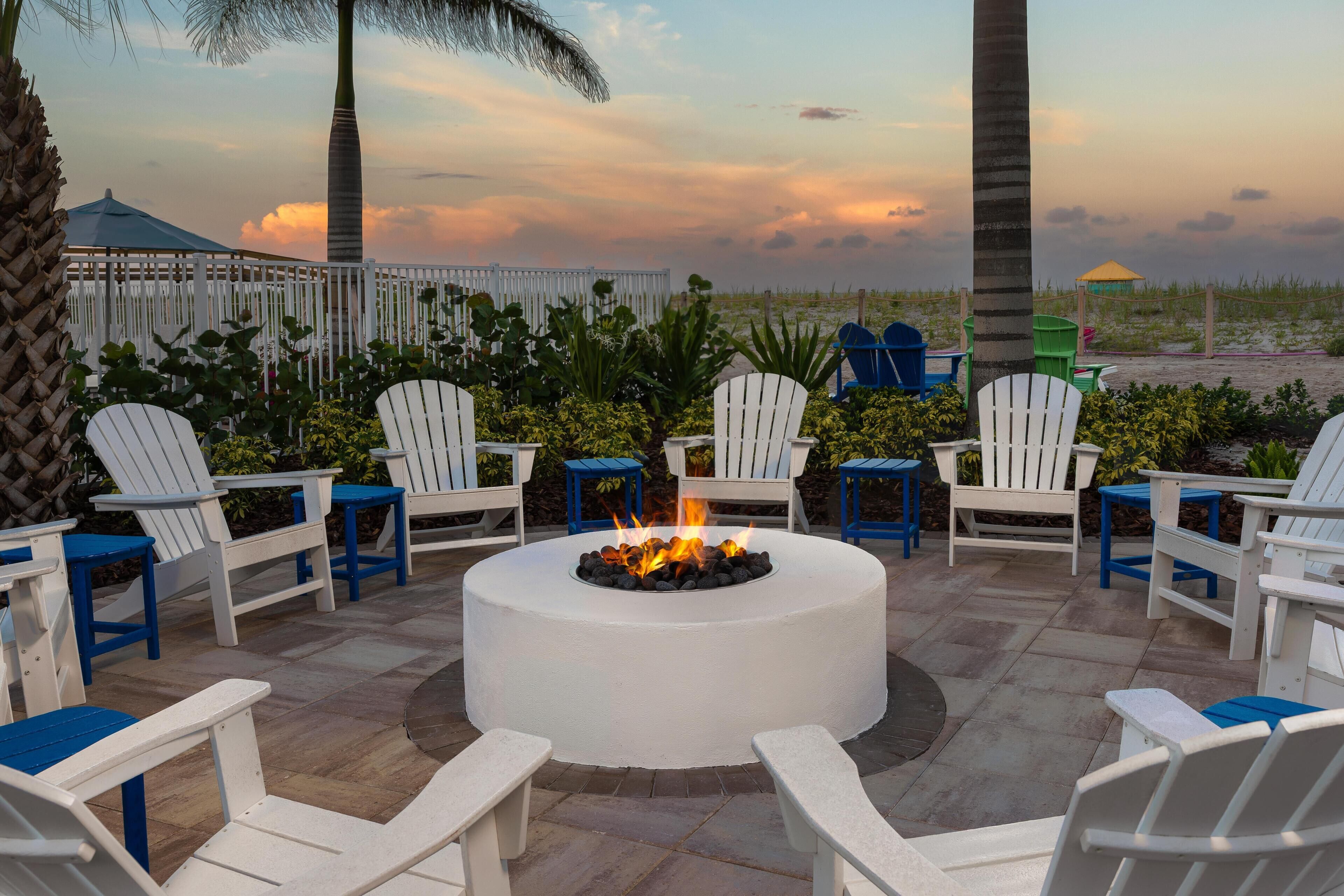 A circular outdoor seating area with a fire pit, surrounded by white and blue Adirondack chairs, and a view of the beach and sky.