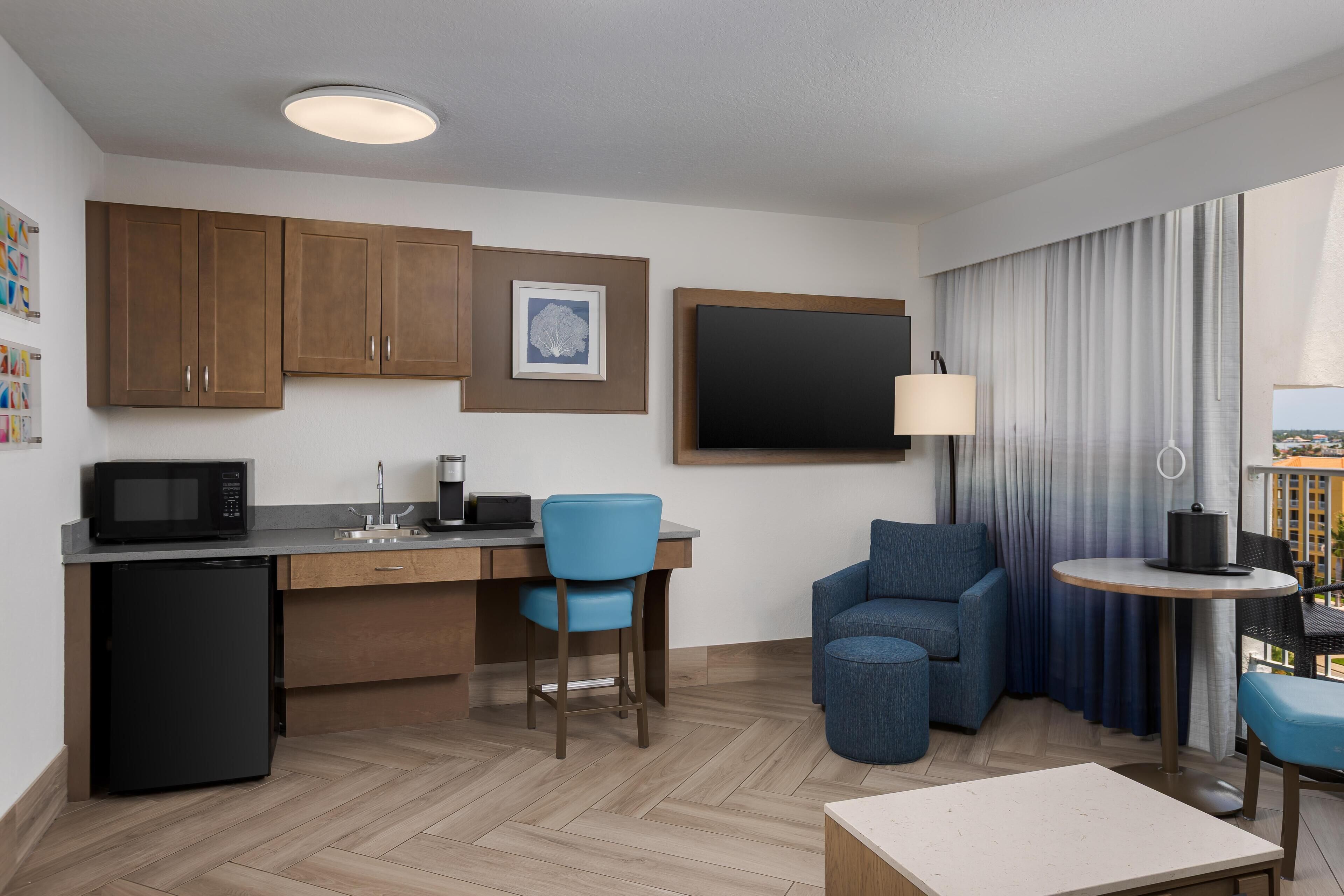 A compact hotel room with a kitchenette, a dining area, a seating area with a sofa and chairs, a small table, and a window with curtains.