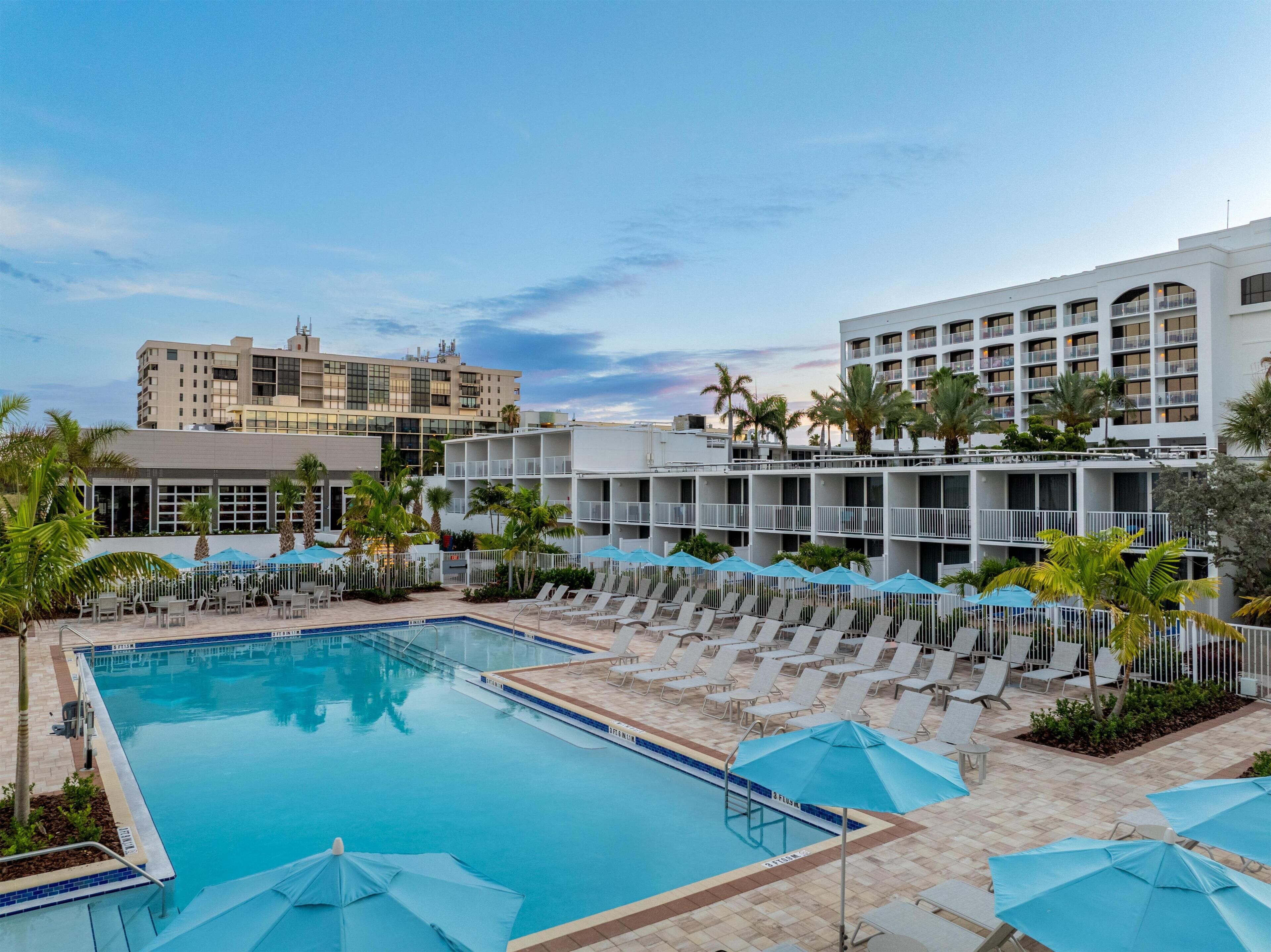 A hotel pool area with sun loungers and umbrellas, surrounded by palm trees and modern buildings.