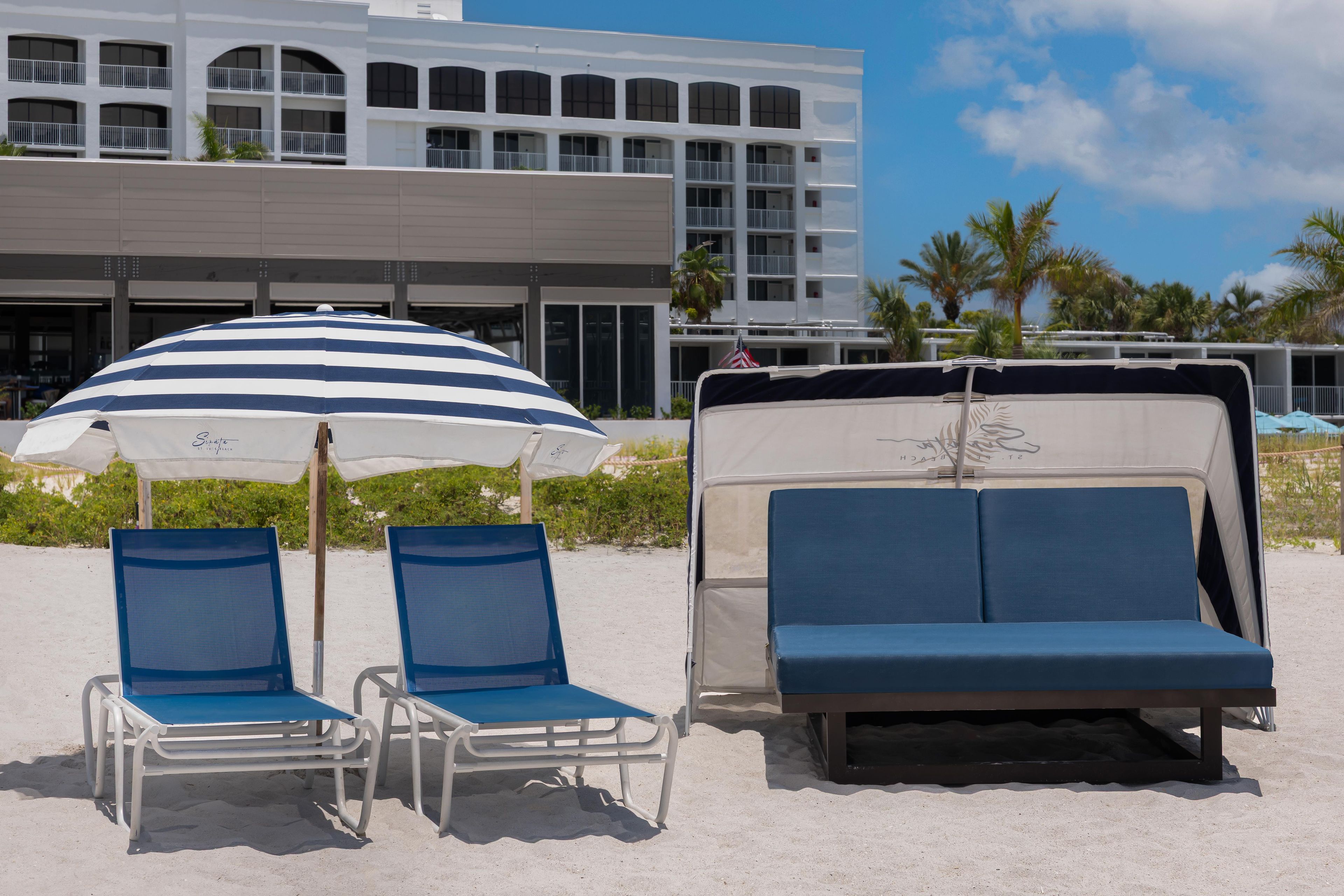 A beachside setting with a white building, two blue lounge chairs, and a striped umbrella.