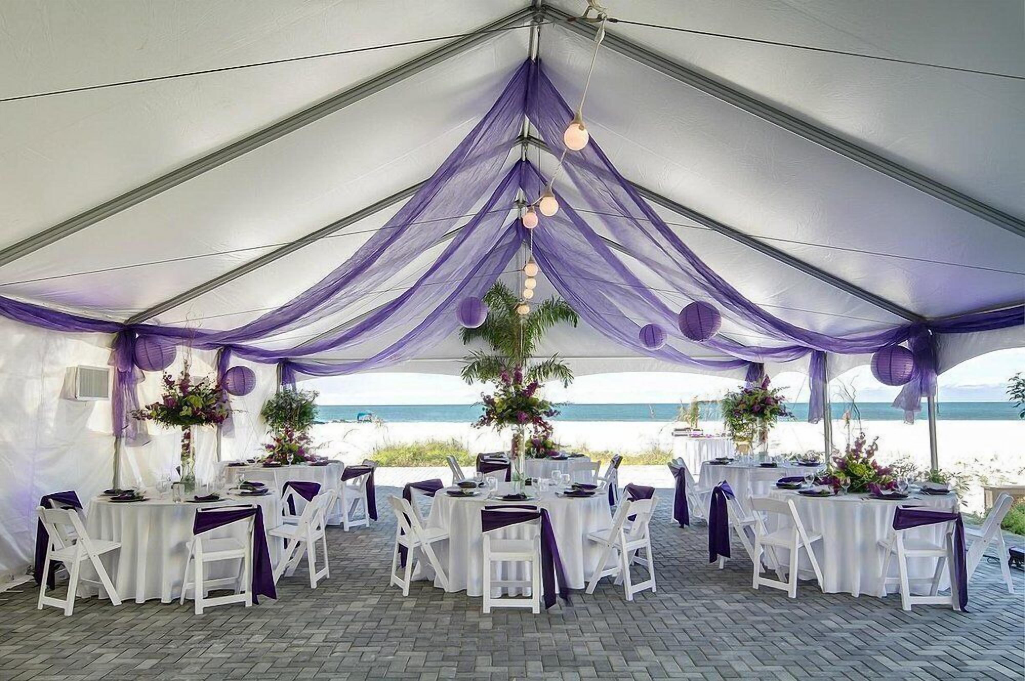 A tented wedding venue with tables and chairs, decorated with purple drapes and floral arrangements, overlooking a beach.