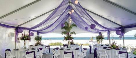 A tented wedding venue with tables and chairs, decorated with purple drapes and floral arrangements, overlooking a beach.