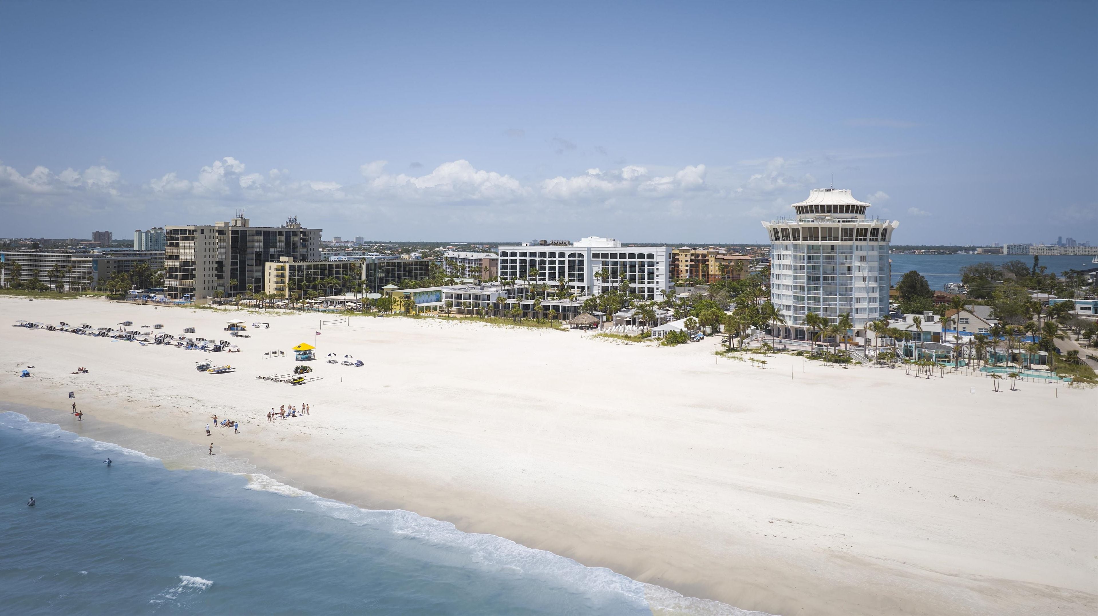 Un resort frente a la playa, con una extensa playa de arena, una alta torre de observación y varios edificios con balcones con vistas al mar.